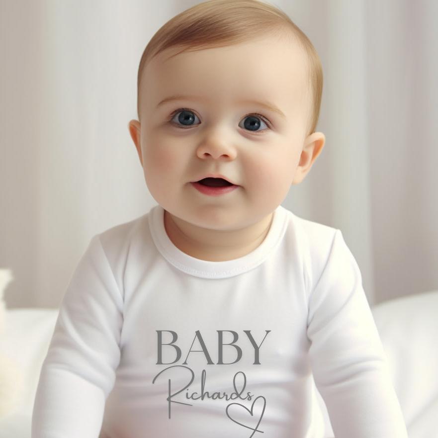 Personalised Surname Heart Baby Outfit featuring a white vest with "BABY Richards" printed in grey, worn by a smiling baby.