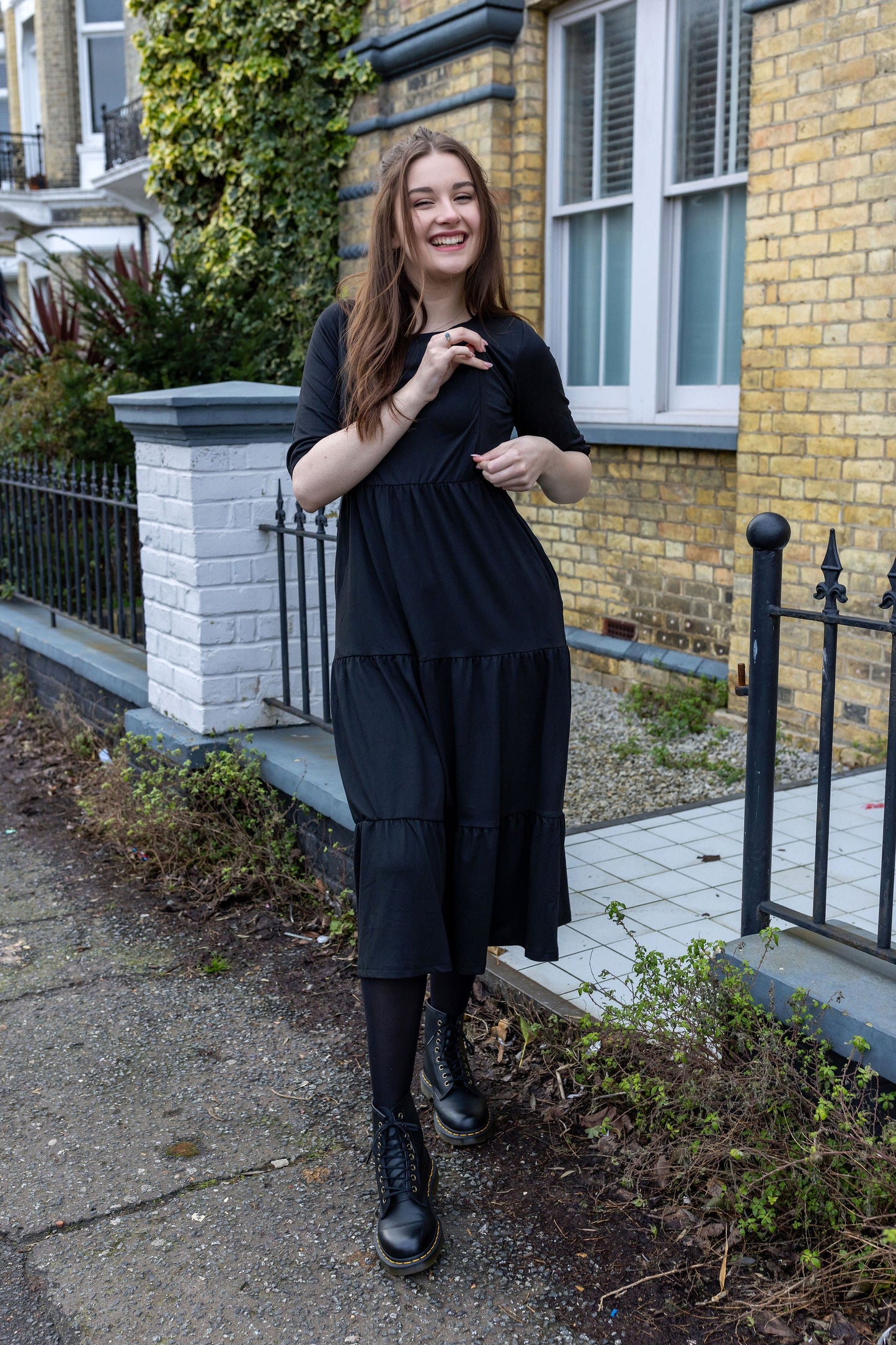 The Midi Breastfeeding Dress - Beth Black Cotton features a smiling woman in a black tiered dress, styled for comfort and nur