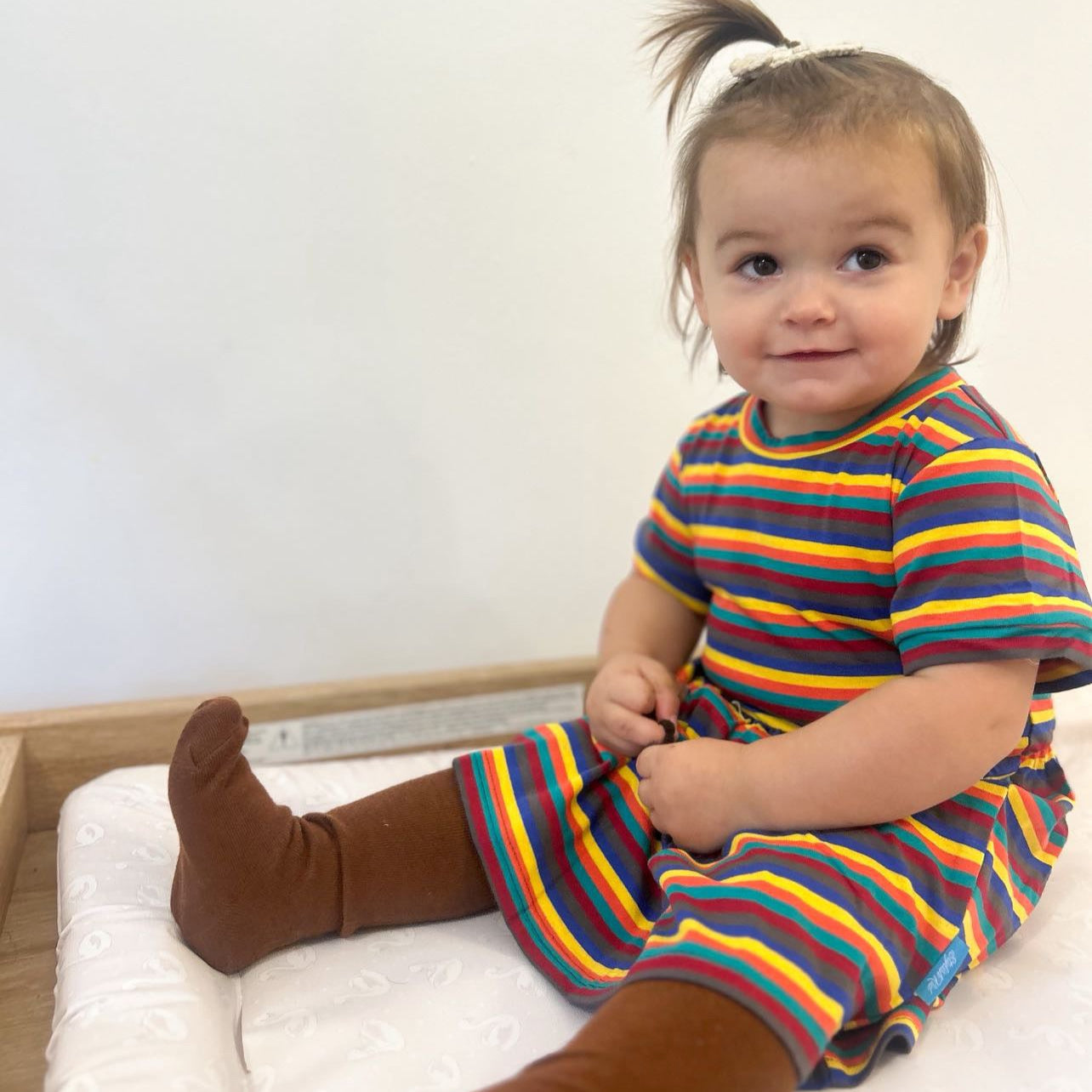 Hazel Rainbow Super Soft Cotton Baby Child Dress featuring colorful stripes, worn by a smiling toddler sitting on a changing 