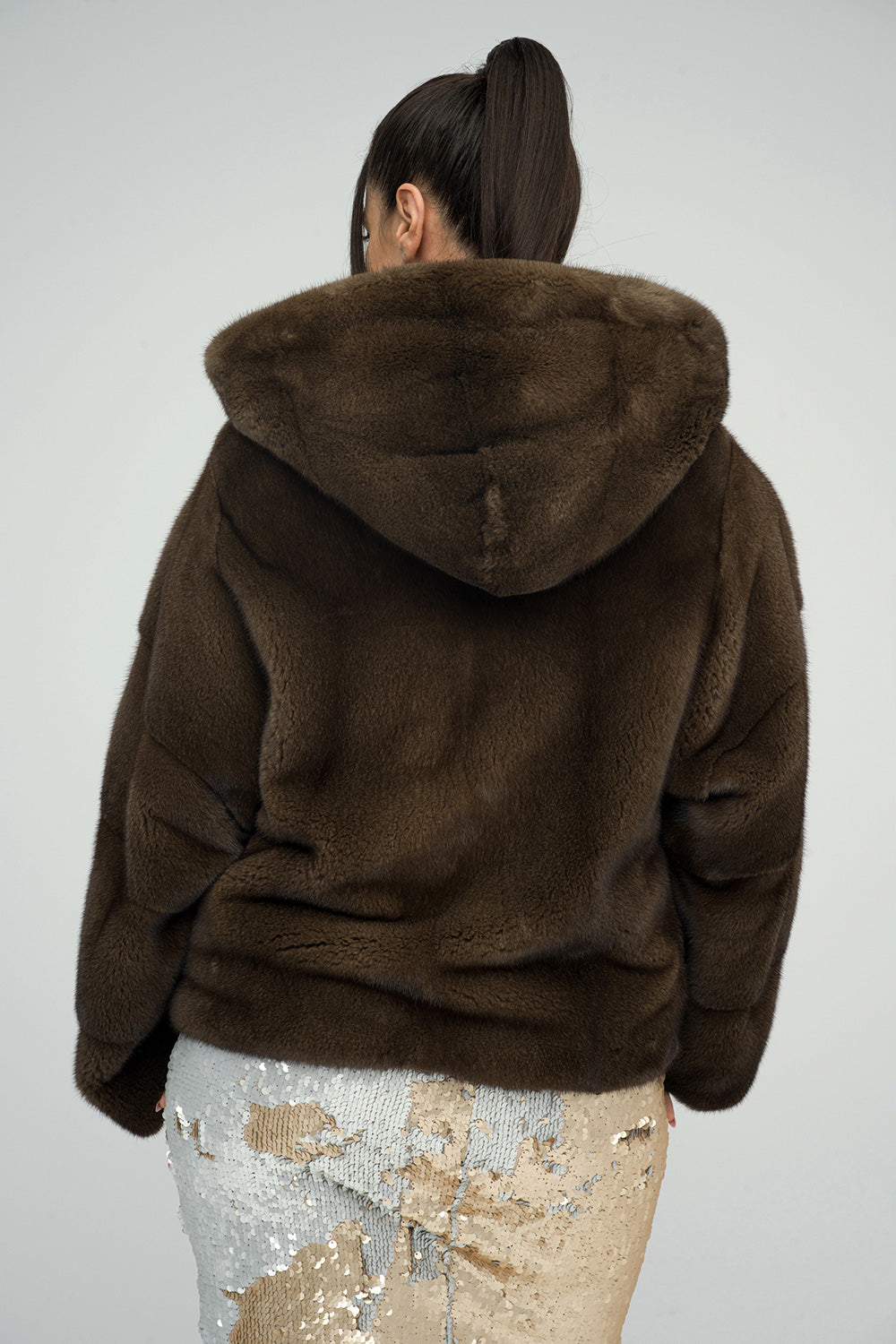 Brown fluffy hooded mink fur coat with a soft texture, featuring a large hood and a relaxed fit, worn over a sequined skirt.