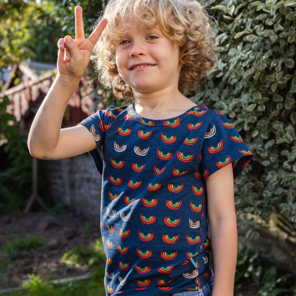 Boob Over the Rainbow Kids top featuring a blue fabric with colorful rainbow patterns, worn by a smiling child outdoors.