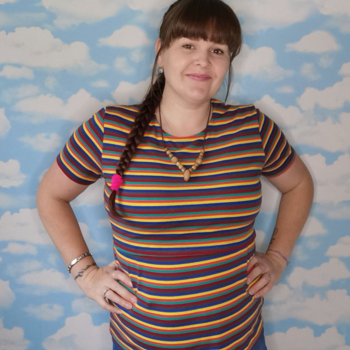 Model wearing a Hazel Stripe Breastfeeding Top with colorful horizontal stripes, smiling against a cloud-patterned background
