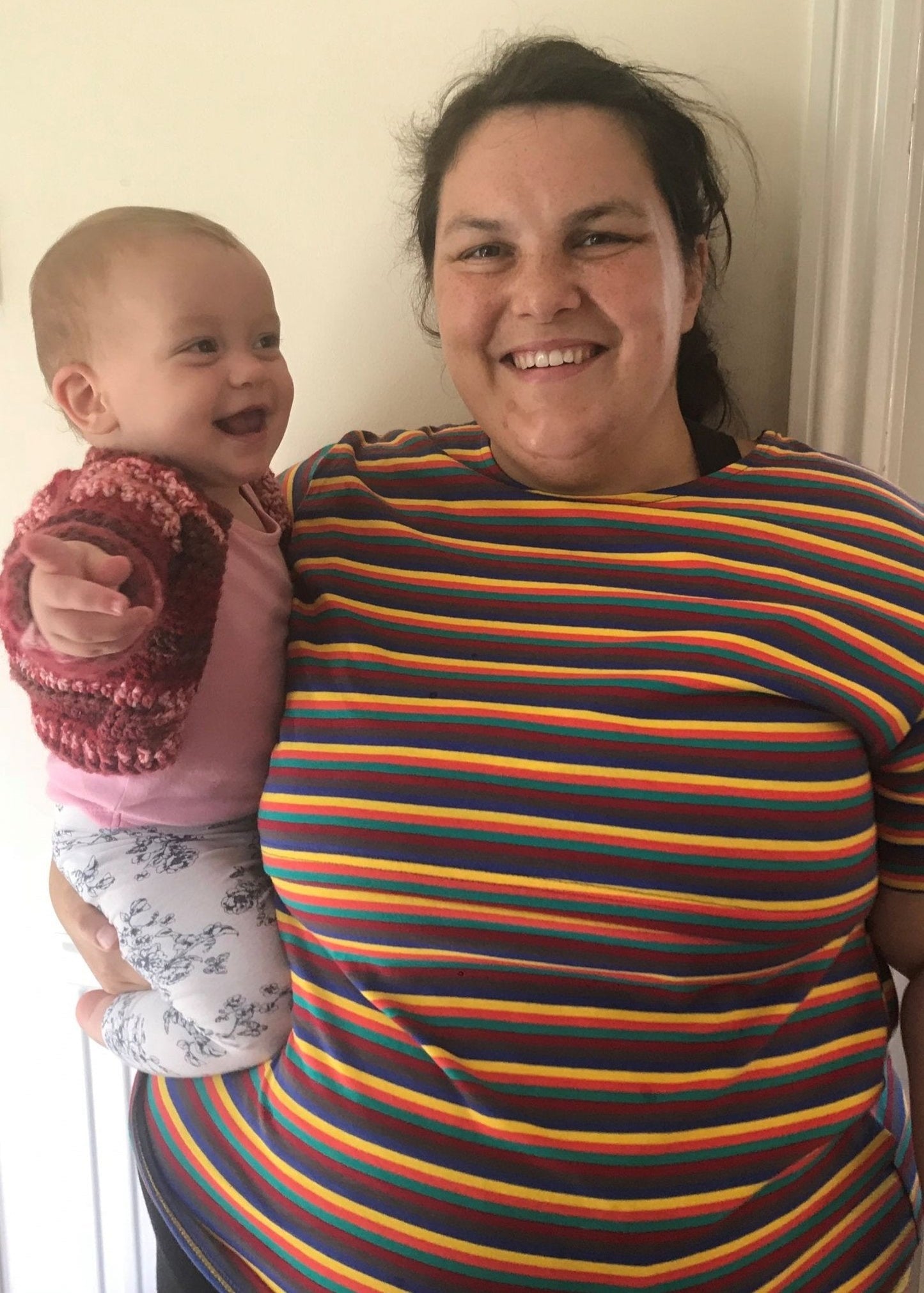 A smiling woman holds a baby while wearing a colorful striped Hazel Stripe Breastfeeding Top.