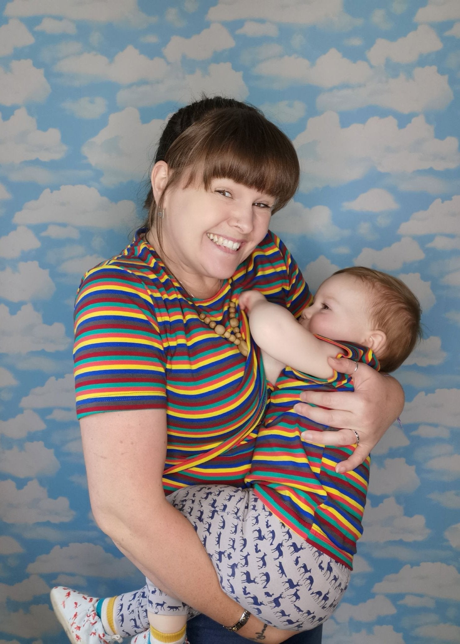 A woman in a striped breastfeeding top holds a baby in a matching striped outfit against a cloud-patterned background.