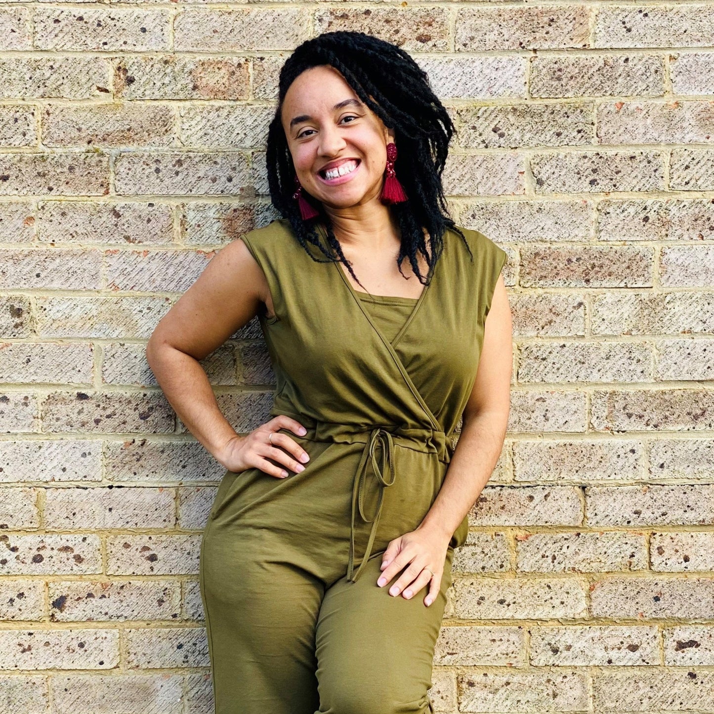 Breastfeeding Jumpsuit in khaki, featuring a wrap design and adjustable tie, modeled by a smiling woman against a brick wall.