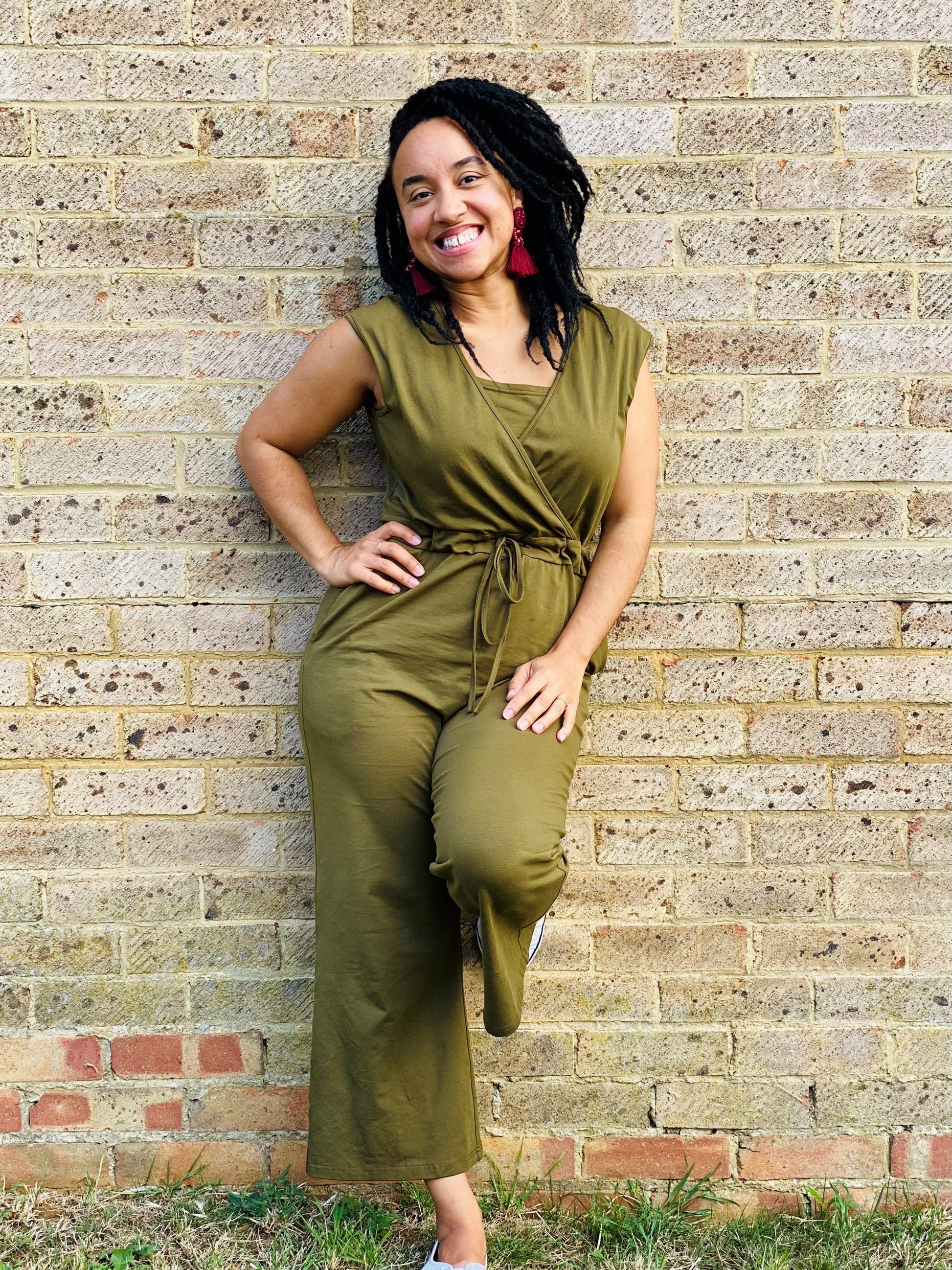 The Breastfeeding Jumpsuit - Kerry Cotton Khaki features a woman in a khaki jumpsuit with a wrap design, standing against a b