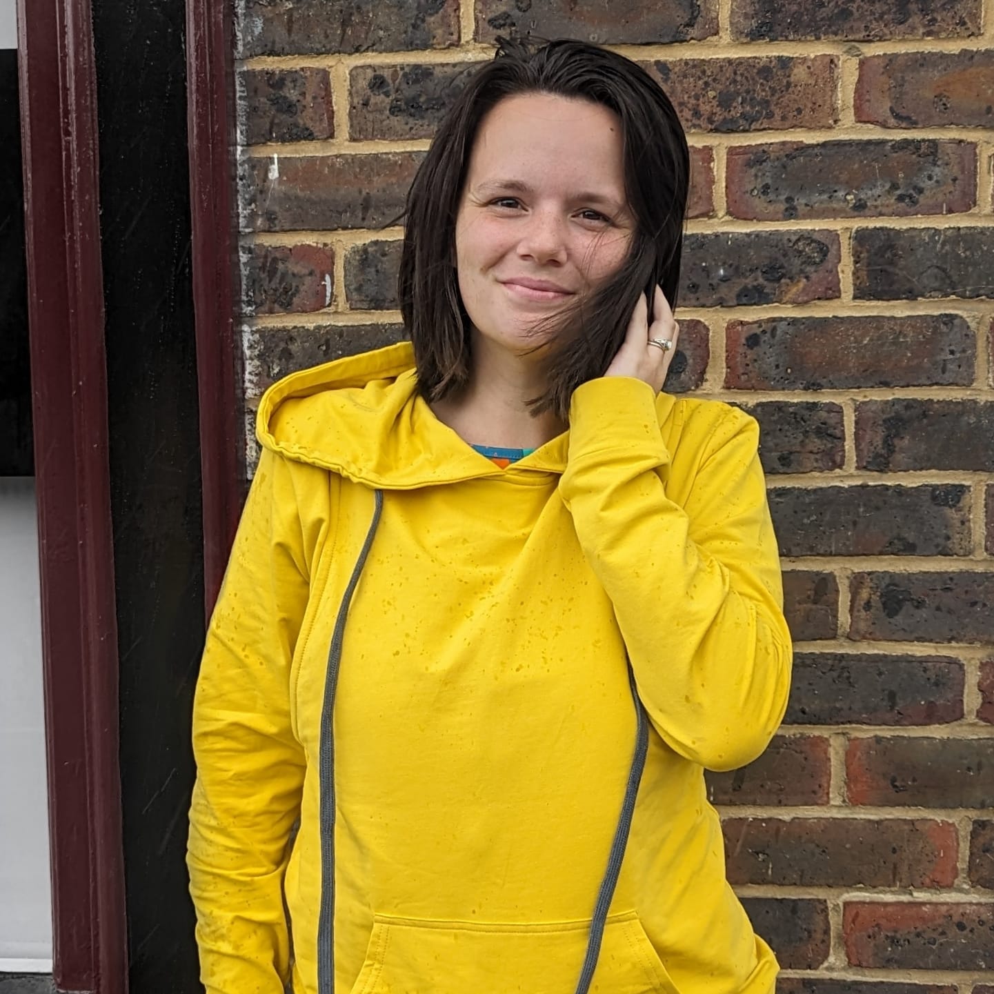 Yellow breastfeeding hoodie worn by a woman, featuring a relaxed fit and drawstring hood, set against a brick wall.
