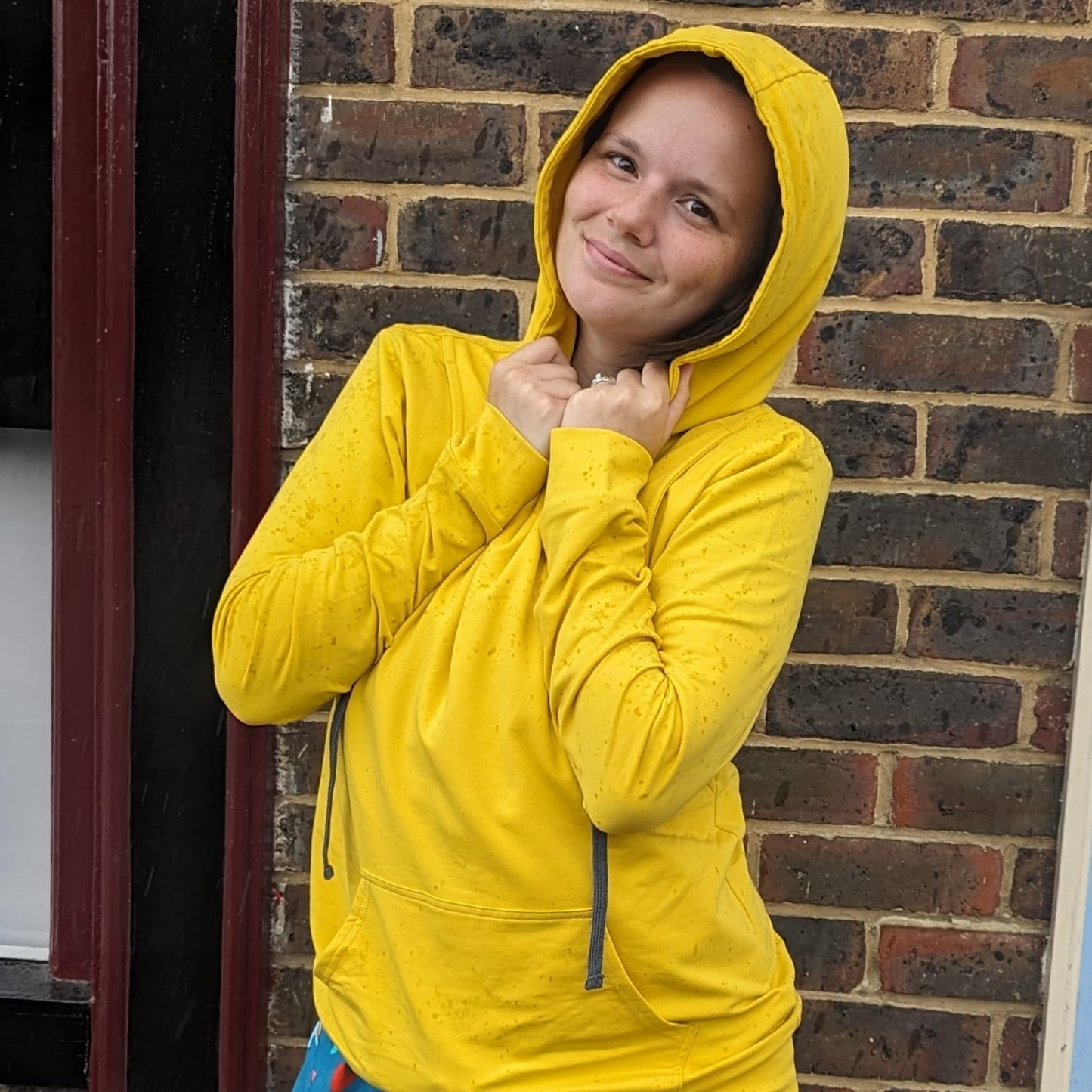 Yellow breastfeeding hoodie featuring a front pocket and adjustable hood, worn by a smiling woman against a brick wall.