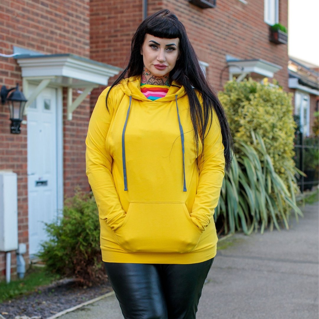 Yellow breastfeeding hoodie with a front pocket, worn by a woman standing outdoors. Features a drawstring hood and relaxed fi